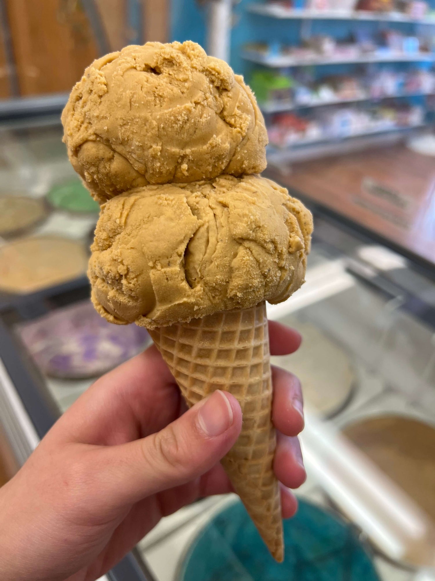A hand holding a double scoop of golden-brown ice cream in a waffle cone inside Liled's Candy Kitchen.