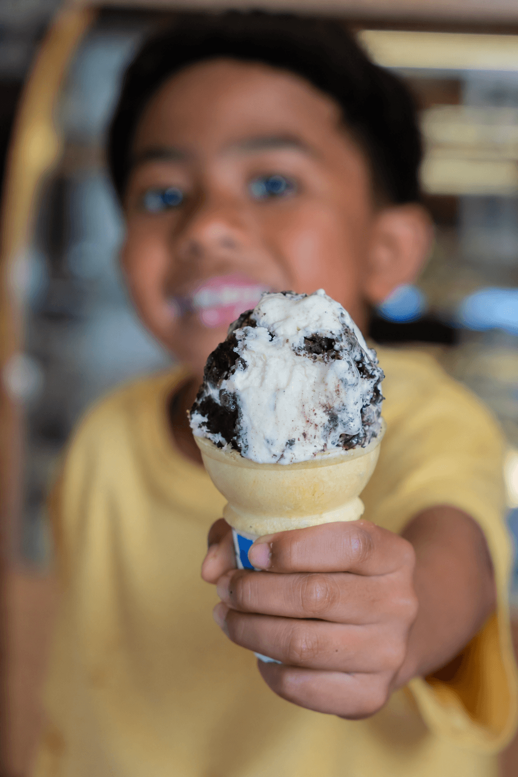 A young boy in a yellow shirt holds a close-up of a vanilla ice cream cone filled with white and dark cookie ice cream, with a blurred background.