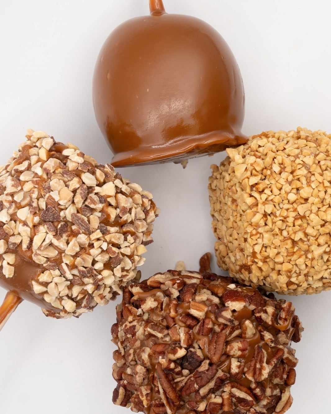 Four decorated chocolate treats on sticks, including one plain milk-chocolate coated piece and three coated pieces topped with chopped nuts in different textures, arranged on a white background.
