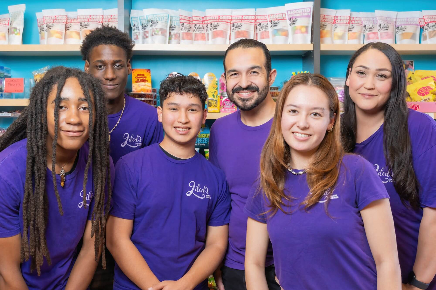 Six Liled's employees wearing matching purple t-shirts with the Liled's logo pose together in front of a store shelf filled with packaged snacks.