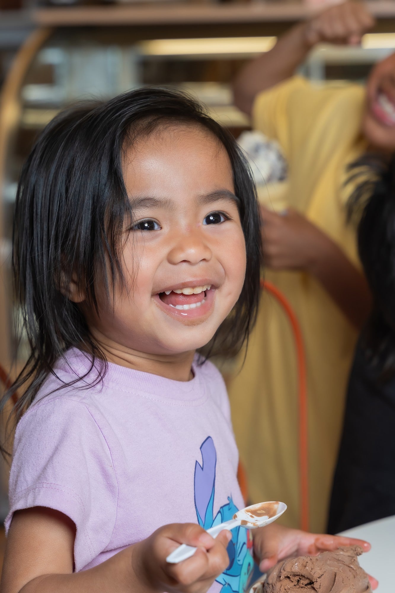 A young child with dark hair smiling while holding a spoonful of chocolate ice cream, wearing a light purple t-shirt with a cartoon graphic. Another person in a yellow shirt is blurred in the background.