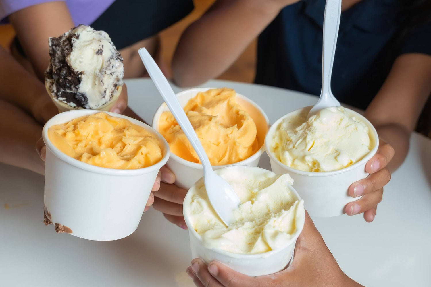 Five paper cups filled with different frozen desserts, including orange custard, vanilla ice cream, and chocolate cookie ice cream, held by hands over a white table. White plastic spoons are in each cup.
