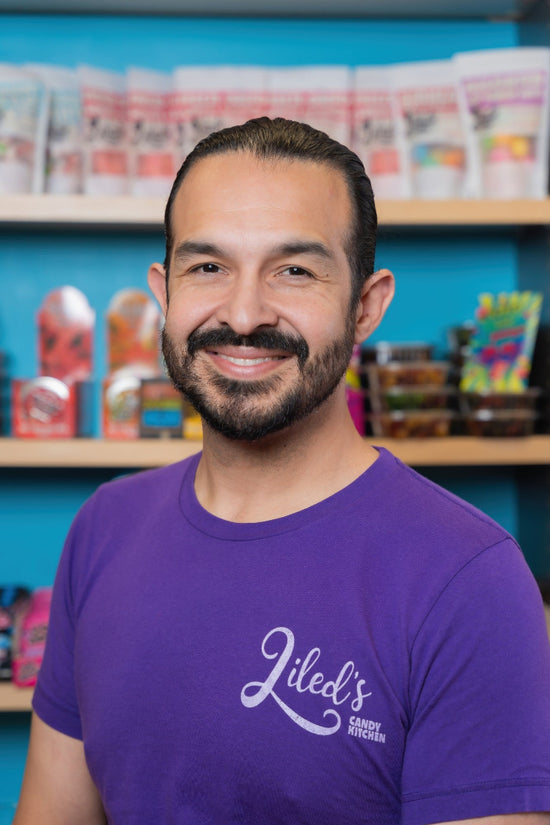 A smiling man with a beard and slicked-back hair wearing a purple t-shirt with 'Liled's Candy Shop' printed in white script, standing in front of shelves filled with colorful packaged snacks.