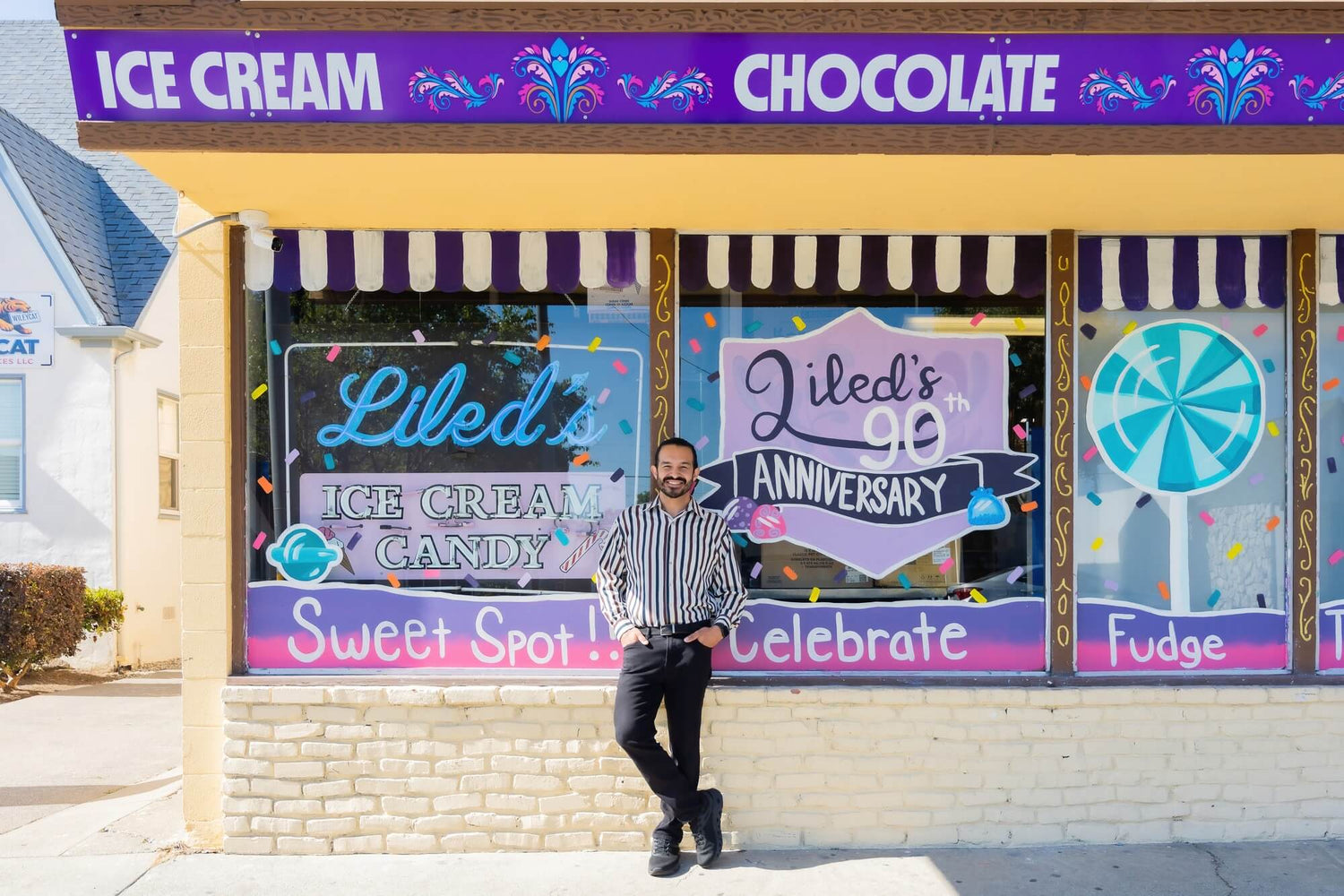 James, the owner of Liled's, stands in front of the colorful Liled's storefront with window decorations reading 'Liled's Ice Cream Candy' and 'Liled's 90th Anniversary.' The building has a yellow facade, a purple sign above, and windows displaying ice cream and chocolate flavors.